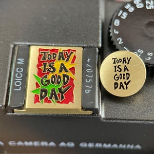May include: A rectangular gold-tone metal pin featuring a red and yellow graphic with the black text "TODAY IS A GOOD DAY" and a circular gold-tone metal pin with the black text "TODAY IS A GOOD DAY" engraved on the surface, both displayed on a camera.