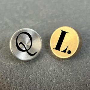 May include: Two round metal buttons, one silver and one gold, each with a black letter. The silver button has a stylized "Q", and the gold button has an "L."