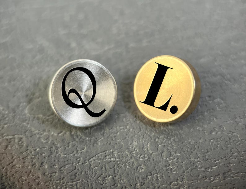 Pode incluir: Dois bot&otilde;es met&aacute;licos circulares, um prateado com a letra Q preta e outro dourado com a letra L preta seguida de um ponto. Ambos os bot&otilde;es possuem uma superf&iacute;cie lisa e plana com acabamento met&aacute;lico e tipografia clara e em negrito.