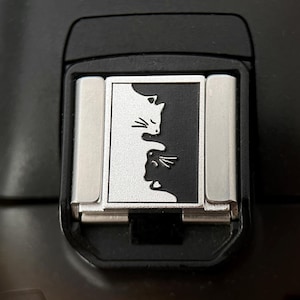 Op de afbeelding: Zwart camera-accessoire met een zilver en zwart kattenontwerp. Het ontwerp toont twee katten silhouetten die tegenover elkaar staan. Het accessoire is rechthoekig met zilveren metalen zijkanten.