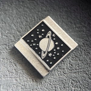May include: A silver-toned metal pin with a black enamel design of a planet with rings and stars.