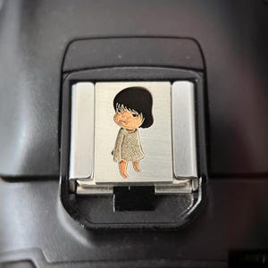 Puede incluir: Cubierta de zapata de cámara plateada con una ilustración de dibujos animados de una niña con cabello negro y expresión neutra. La niña lleva un vestido de color claro y tiene los pies naranjas. La cubierta está sobre un cuerpo de cámara negro.