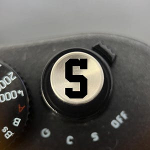 Pode incluir: Um bot&atilde;o de disparo de c&acirc;mara circular prateado com uma letra S preta proeminente no centro. O bot&atilde;o est&aacute; inserido num corpo de c&acirc;mara preto com um seletor de velocidade do obturador &agrave; esquerda, exibindo os n&uacute;meros 1000, 2000 e 4000.