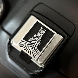May include: Close-up of a black camera accessory with a silver and black zebra design. The zebra is depicted in a stylized, minimalist form. The accessory is rectangular with rounded corners and a metallic frame.