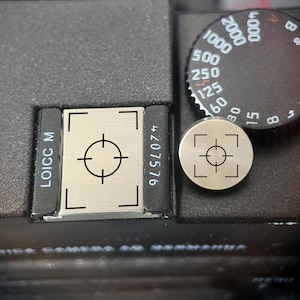 Pode incluir: Uma tampa retangular de sapata de flash em metal prateado e um botão disparador circular em metal prateado, ambos com um gráfico de mira preto. A peça retangular está gravada com as inscrições LOICC M e 4207576.