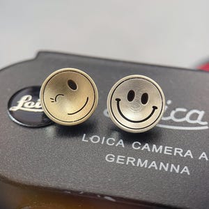 May include: A pair of round stud earrings featuring smiley faces. One earring is gold-toned and winking, the other is silver-toned and smiling. The earrings are on a dark surface with the text "Leica Camera AG Germanna".