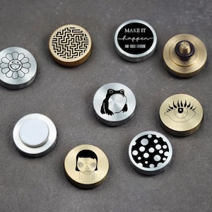 May include: A collection of nine circular metal buttons or pins featuring various designs including a flower, a maze, a frog, an eye, a person, polka dots, and one with the text "MAKE IT happen AND SHOCK EVERYONE" in black and white.