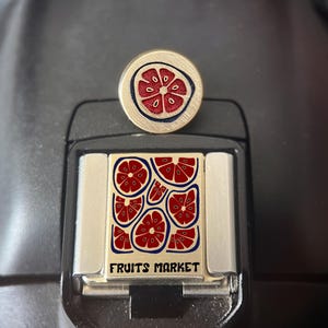 Puede incluir: Un pin metálico circular con un diseño de rodaja de pomelo rojo y una placa metálica rectangular con un patrón repetitivo de rodajas de pomelo rojo y el texto FRUITS MARKET en letras negras, ambos con un acabado en tono dorado cepillado.