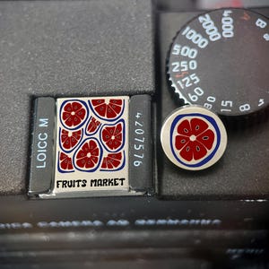 May include: A black camera body with a small rectangular tag displaying "FRUITS MARKET" and a design of red citrus fruit slices. A round pin with a similar citrus fruit design is also visible. The camera's settings dial is also in view.