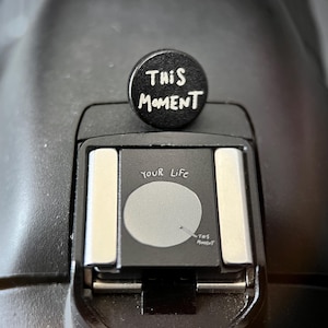 Könnte beinhalten: Eine schwarze Blitzschuhabdeckung für die Kamera mit dem weißen Text "THIS MOMENT". Der Blitzschuh der Kamera trägt den weißen Text "YOUR LIFE" und "THIS MOMENT". Die Kamera ist schwarz.