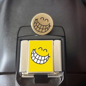 Può includere: Una spilla metallica circolare con un volto sorridente inciso in nero, occhi chiusi e un ampio sorriso a denti stretti, posizionata sopra un copri slitta per flash rettangolare che mostra una grafica gialla abbinata dello stesso volto.