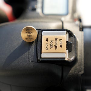 May include: A close-up of a black camera with a gold-colored round button and a rectangular card. The button and card both have the text "your image. logo. or text". The camera is in focus.