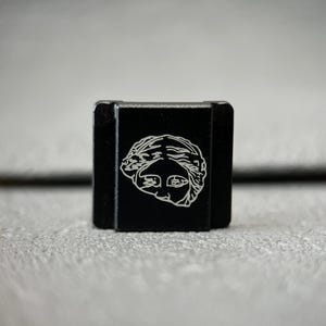 Puede incluir: Pequeño objeto rectangular negro con una ilustración blanca estilizada de una cara en la parte delantera. El objeto parece ser de metal y tiene un acabado mate. El fondo es de color neutro borroso.