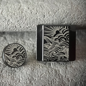 May include: A black, round knob and a black, rectangular object with a sun and wave design in white. The knob is circular, while the rectangular object has a square shape. Both items feature a detailed, engraved illustration.