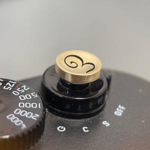 Pode incluir: Um bot&atilde;o de disparo circular em tom de lat&atilde;o com uma grava&ccedil;&atilde;o estilizada do n&uacute;mero 3, montado na parte superior preta de uma c&acirc;mara. A c&acirc;mara possui um disco preto com marca&ccedil;&otilde;es brancas de velocidade do obturador: 30, 60, 125, 250, 500, 1000, 2000, 4000 e B.
