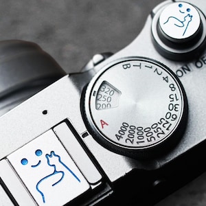 May include: A silver camera with a black dial that has numbers and letters on it. The dial has a red "A" and the numbers 4000, 2000, 1000, 500, 250, 125, 60, 30, 15, 8, 2, 4, 7, T, B. The camera also has a black button with the words "ON OFF" on it.