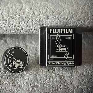 Puede incluir: Tarjeta SD Fujifilm negra y un pin redondo con un diseño de escena de parque. La tarjeta SD presenta el logotipo de Fujifilm, el indicador REC y el texto "Street Photographer". El pin tiene un diseño de árbol, banco y pájaro.