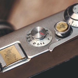 May include: Close-up of a vintage silver camera with a gold-colored accent featuring a skeleton design. The camera has multiple dials and settings, including a shutter speed dial and a film advance lever. The skeleton design is etched into the metal.