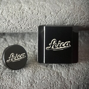 Op de afbeelding: Twee zwarte camera-accessoires, een ronde lensdop en een rechthoekige doos, beide met het Leica-logo in witte letters. Ze liggen op een gestructureerd grijs oppervlak.