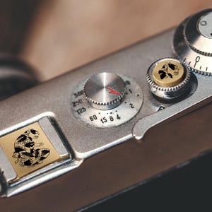 Op de afbeelding: Close-up van een vintage zilveren camera met zwarte en gouden accenten. De camera heeft een reeks draaiknoppen en knoppen, waaronder een snelheidsregelaar met rode markeringen en een kleine, ronde knop met een schedelontwerp. Een rechthoekige gouden plaat met een zwart-witte illustratie is ook zichtbaar.