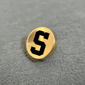 Pode incluir: Um bot&atilde;o met&aacute;lico circular de tom dourado com uma letra S preta em negrito e estilo bloco impressa no centro. O bot&atilde;o tem um acabamento met&aacute;lico liso com uma borda ligeiramente elevada, apresentando um design claro e simples para roupas.