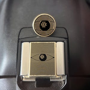 Könnte beinhalten: Ein runder Ansteckpin aus goldfarbenem Metall und eine rechteckige Blitzschuhabdeckung aus goldfarbenem Metall, beide mit einem schwarz-goldenen Linienmuster eines Auges und dem Text EYE SEE YOU in der Pupille.
