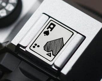 May include: A close-up of a silver camera with a black ace of spades symbol on the top. The symbol is surrounded by a white border.
