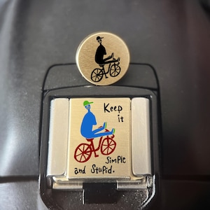 May include: A round gold-toned pin featuring a black silhouette of a person riding a bicycle, paired with a rectangular gold-toned charm displaying a blue person on a red bicycle with the text Keep it simple and Stupid in black lettering.