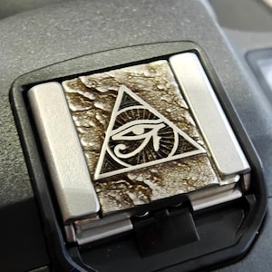 May include: A silver camera hot shoe cover with a textured, metallic finish. The cover features a raised design of an eye inside a triangle.