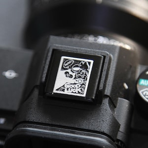 May include: A black camera with a silver metal plate on the top. The plate has a cartoon graphic of a dog with the words "Good Luck" printed on it.