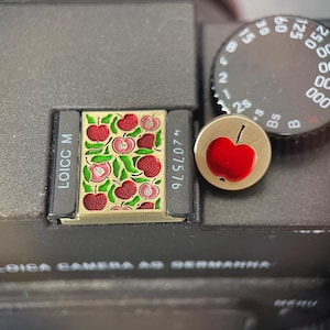 Puede incluir: Una tapa de zapata rectangular de metal con un patrón repetitivo de manzanas rojas y hojas verdes, junto a un botón disparador circular de metal con una incrustación de manzana roja, sobre un cuerpo de cámara negro con texto visible.