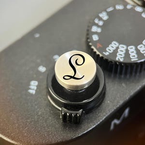 Pode incluir: Um bot&atilde;o disparador de c&acirc;mara circular de tom prateado com uma letra L preta gravada em fonte cursiva. O bot&atilde;o est&aacute; montado no corpo preto de uma c&acirc;mara ao lado de um seletor de velocidade do obturador preto com marca&ccedil;&otilde;es num&eacute;ricas brancas.