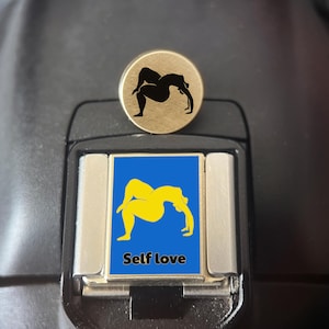 Peut inclure: Une épinglette ronde en métal doré avec une silhouette noire d'une personne en posture de yoga, placée au-dessus d'une breloque rectangulaire en métal argenté avec un fond bleu, une silhouette jaune en posture de yoga et le texte Self love.