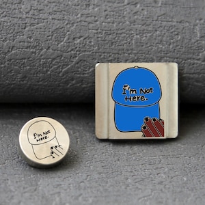 May include: A circular metal pin and a square metal pin, both featuring a graphic of a blue baseball cap with the text "I'm Not Here." and a hand reaching toward the cap. The circular pin has a black line drawing, while the square pin is coloured.