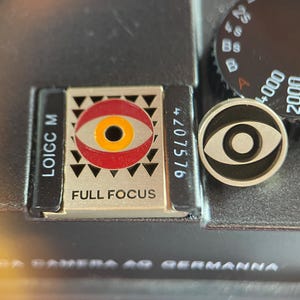 May include: A rectangular metal pin featuring a red and yellow eye graphic with the text FULL FOCUS and LOIGC M, alongside a circular black and silver pin with an eye icon, both resting on a black camera body with a shutter speed dial.