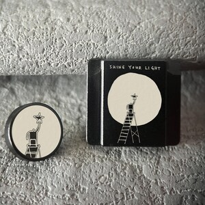 May include: A black and white illustration of a person on a ladder reaching for a star, with the text "Shine Your Light." The design is featured on a round pin and a small black box.