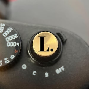Pode incluir: Um bot&atilde;o disparador de c&acirc;mara circular de cor dourada com uma letra L preta e um ponto. O bot&atilde;o est&aacute; montado num corpo de c&acirc;mara preto ao lado de um seletor de velocidade do obturador com n&uacute;meros brancos de 2s a 4000 e defini&ccedil;&otilde;es B.