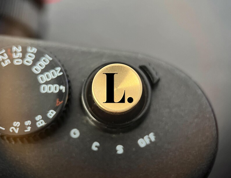 Pode incluir: Um bot&atilde;o disparador de c&acirc;mara circular de cor dourada com uma letra L preta e um ponto. O bot&atilde;o est&aacute; montado num corpo de c&acirc;mara preto ao lado de um seletor de velocidade do obturador com n&uacute;meros brancos de 2s a 4000 e defini&ccedil;&otilde;es B.