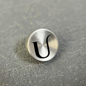 Pode incluir: Um bot&atilde;o met&aacute;lico circular de tom prateado com acabamento escovado e uma letra U preta gravada no centro. O bot&atilde;o tem uma superf&iacute;cie lisa e ligeiramente abaulada com uma textura radial distinta que irradia a partir do centro.