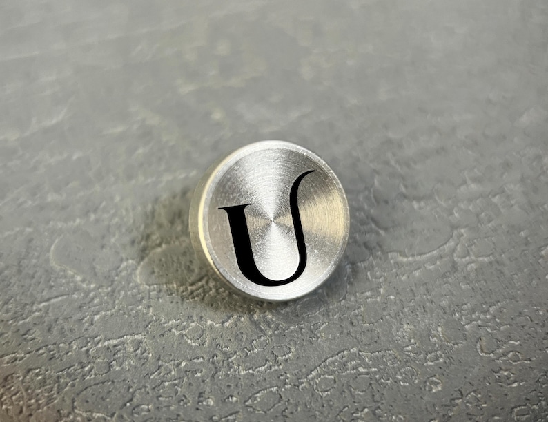 Pode incluir: Um bot&atilde;o met&aacute;lico circular de tom prateado com acabamento escovado e uma letra U preta gravada no centro. O bot&atilde;o tem uma superf&iacute;cie lisa e ligeiramente abaulada com uma textura radial distinta que irradia a partir do centro.