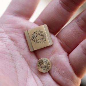 May include: Two small, gold-toned metal objects, one square and one round, each with a black engraved image of a classical female face. The objects are held in a hand, showing their size and detail. The square object has a rectangular shape.
