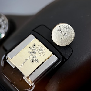 Op de afbeelding: Een goudkleurig metalen accessoire met een rozenillustratie. Het ontwerp is te zien op een rechthoekig stuk en een cirkelvormig stuk, beide met een zwarte rozenstengel en bladeren. De stukken zijn geplaatst op een zwarte camera.