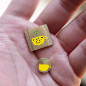 Pode incluir: Dois pequenos pinos bege. Um é quadrado, o outro redondo. Ambos apresentam um desenho de chávena amarela com um rebento verde e o texto "Do what makes your soul SHINE!"
