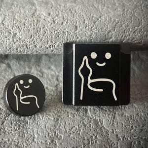 Könnte beinhalten: Zwei schwarze Dekorationsgegenstände mit weißem Grafikdesign. Einer ist rund, der andere quadratisch. Das Design zeigt ein lächelndes Gesicht und eine stilisierte Figur. Die Objekte befinden sich vor einem strukturierten grauen Hintergrund.