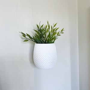 Puede incluir: Maceta de cerámica blanca con un diseño de espiral, colgada de una pared blanca. La maceta está llena de plantas verdes de hoja.