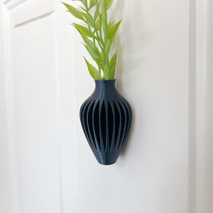 May include: A dark blue, 3D-printed vase with vertical, curved cutouts. The vase holds a bundle of green, leafy stems. The vase is mounted on a white door.