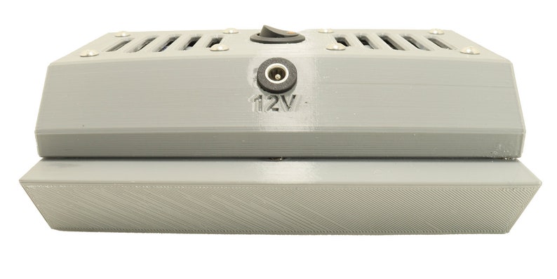 May include: A grey, rectangular, 3D printed box with a power cord and a switch on the top. The box has a vent on the top and a label that says "12V".