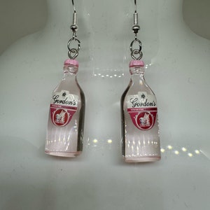Könnte beinhalten: Ein Paar silberne Ohrringe mit rosa Flaschenanhängern. Die Flaschen haben ein klares Glas-Design und tragen ein Etikett mit der Aufschrift "Gordon's Gin".