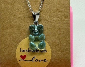 May include: A silver chain necklace with a light blue gummy bear pendant. The pendant is translucent and has a classic gummy bear shape. The necklace is displayed on a brown card with a sticker that says "handmade with love."