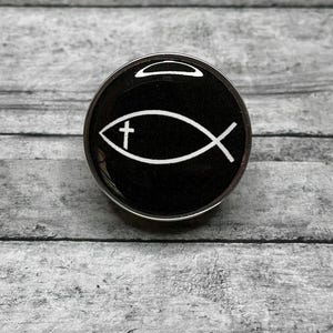 May include: A round, black button with a silver rim. The button features a white Ichthys symbol, a Christian symbol of a fish with a cross inside. The button is set against a gray wooden background.